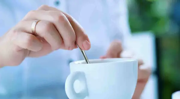 Mano con anello mescola il caffè in una tazza bianca. La scelta è sempre la stessa: caffè con o senza zucchero.