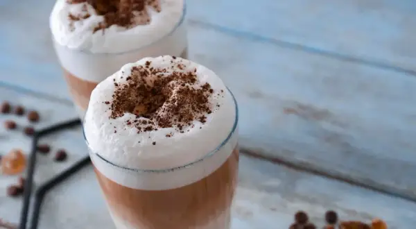 Due bicchieri di latte macchiato freddo con schiuma soffice e cacao in polvere, su un tavolo di legno azzurro.