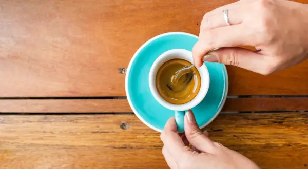 Mani che mescolano un caffè, nonostante si dica che il caffè è cancerogeno, la tazzina turchese su un tavolo di legno è invitante.
