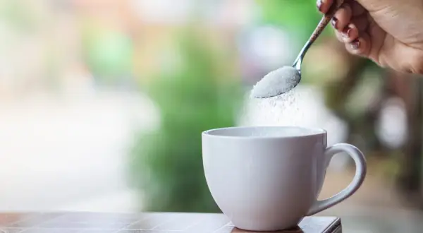 Mano con cucchiaino che versa zucchero in una tazza bianca, pronta per il caffè.