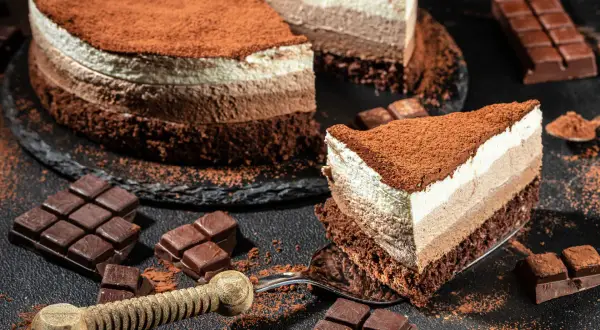 Una golosa cheesecake cappuccino a tre strati, cosparsa di cacao, con pezzi di cioccolato e una fetta servita su una forchetta.