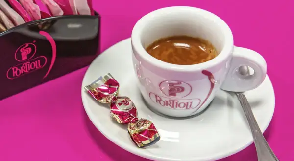 Una tazzina di crema al caffè espresso, con tre caramelle incartate e un porta-bustine Portioli su sfondo fucsia.