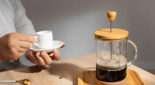 Un uomo tiene una tazzina di caffè mentre una french press caffè è pronta per essere servita.