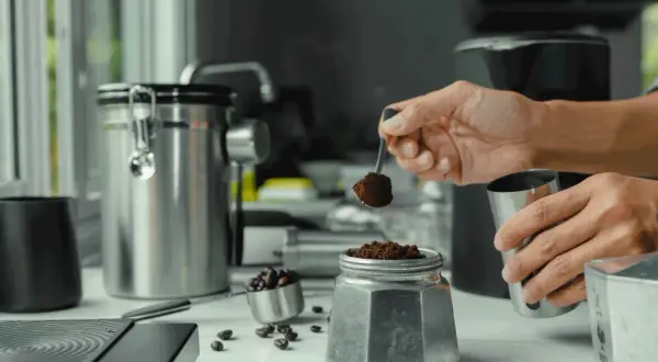 Mano aggiunge caffè macinato a una moka, per il suo primo utilizzo. Caffè in grani e contenitori in acciaio completano la scena.