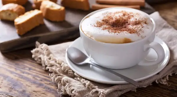 Un invitante cappuccino caldo, spolverato di cacao, accanto a biscotti croccanti e cannella su un tavolo di legno.