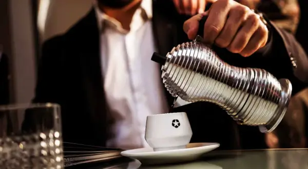 Un uomo in giacca e camicia versa caffè da un bricco argentato in una tazzina bianca. La macinatura caffè più fine garantisce un aroma intenso.