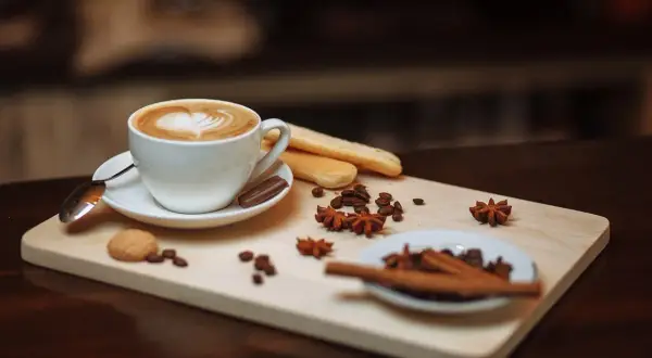 Un cappuccino macchiato con schiuma a forma di cuore, biscotti, chicchi di caffè e spezie su un tagliere di legno.
