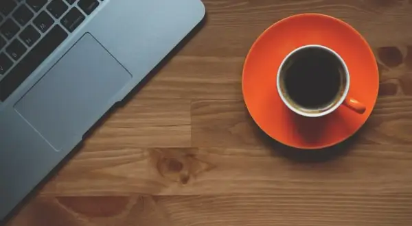 Tazza di caffè nero su piattino arancione accanto a un laptop. Scopri quando bere il caffè per dimagrire.