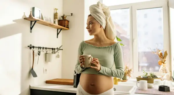 Donna incinta con asciugamano in testa beve una tisana in cucina, un momento di relax in gravidanza.