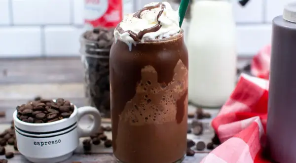 Un delizioso Frappuccino Java Chip, con panna montata e sciroppo al cioccolato. Sullo sfondo, chicchi di caffè, gocce di cioccolato e latte, tutti ingredienti per prepararlo.