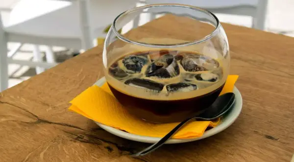 Un bicchiere di caffè freddo con ghiaccio, servito su un piattino con tovagliolo giallo e cucchiaino. Un rinfrescante caffè pugliese per una giornata calda.