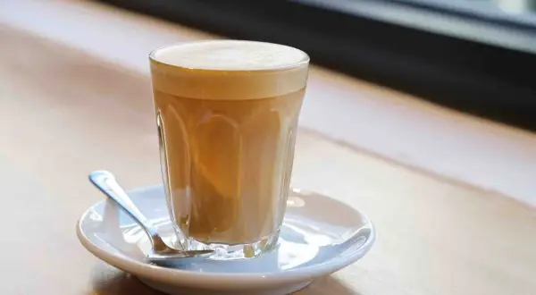 Un bicchiere di cafe latte con schiuma cremosa, servito su un piattino con un cucchiaino, su un tavolo di legno.
