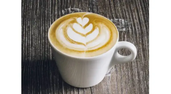 Un flat latte con schiuma artistica a forma di cuore e petali, servito in una tazza bianca su un tavolo di legno scuro.