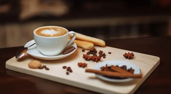 Un delizioso caffè fatto in casa con schiuma a forma di cuore, biscotti, chicchi di caffè e spezie su un tagliere di legno.