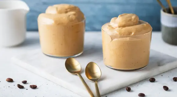 Due bicchieri di soffice crema al caffè senza panna, preparata con caffè moka, su un tagliere di marmo con chicchi di caffè e cucchiaini dorati.