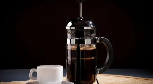 Una tazzina bianca accanto a una caffettiera francese piena di caffè scuro, pronta per essere gustata.