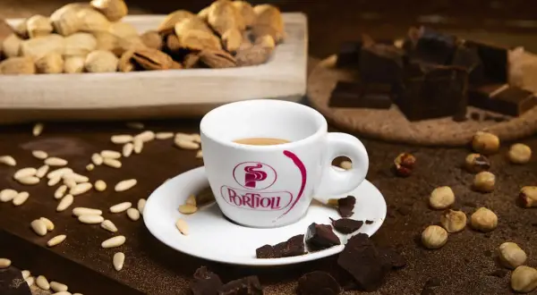 Tazza di caffè fumante, circondata da pezzi di cioccolato fondente, nocciole e pinoli. Un'invitante combinazione di caffè e cioccolato.