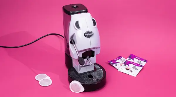 Didiesse Frog espresso machine with coffee pods and Lavazza Gran Espresso Intenso packets on a pink background.