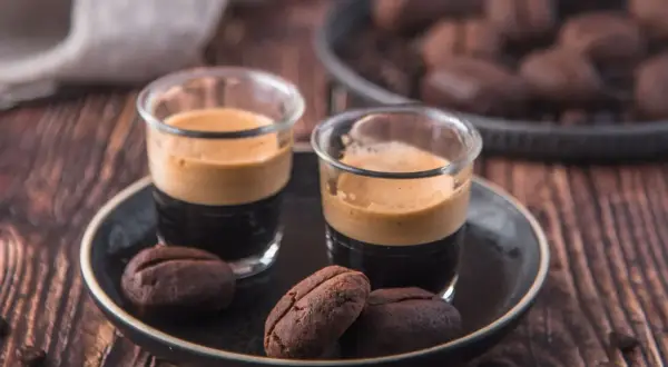 Due bicchierini di espresso con crema e biscotti al caffè moka a forma di chicco, su un piatto scuro.