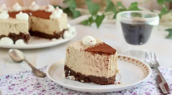Una fetta di cheesecake al caffè senza gelatina, con base croccante al cacao e decorata con panna e cacao in polvere.