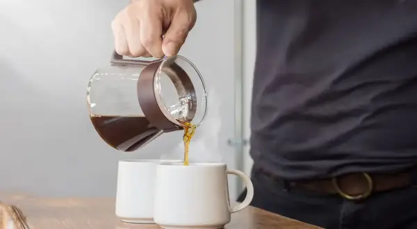 Un uomo versa caffè americano con latte in due tazze bianche su un tavolo di legno.
