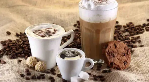 Tre tazze di caffè, una con panna e cacao, un'altra con schiuma e un bicchiere alto di latte cappuccino, accompagnate da biscotti e chicchi di caffè.