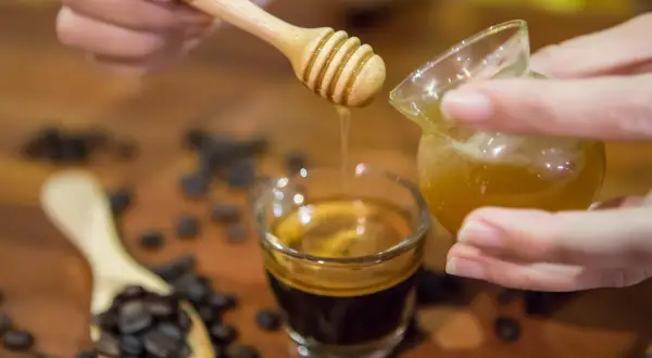 Gocce dorate di miele scendono su un caffè espresso. Chicchi di caffè tostati e un cucchiaino di legno completano la scena.