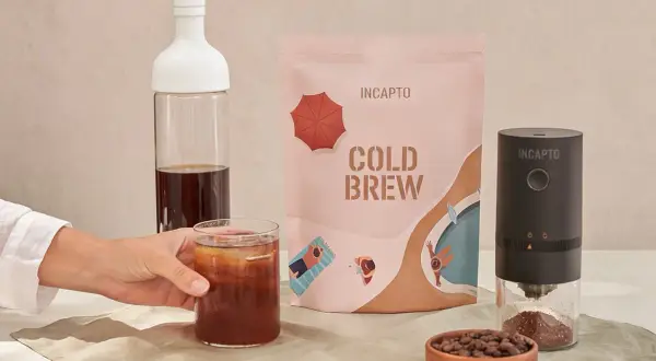 Un bicchiere di caffè freddo in bottiglia, pronto per essere gustato. Accanto, una confezione di cold brew e un macinacaffè.