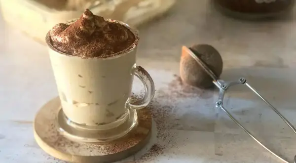 Deliziosa cremina caffè in una tazza di vetro, spolverata di cacao. Accanto, un colino per dolci e un barattolo di caffè.