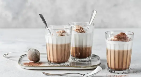 Tre bicchieri di cappuccino con cioccolato, guarniti con panna e cacao, su un vassoio con colino.