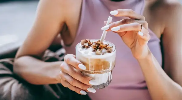 Mani con unghie bianche sorreggono un bicchiere di delizioso **caffè freddo** con topping croccante.