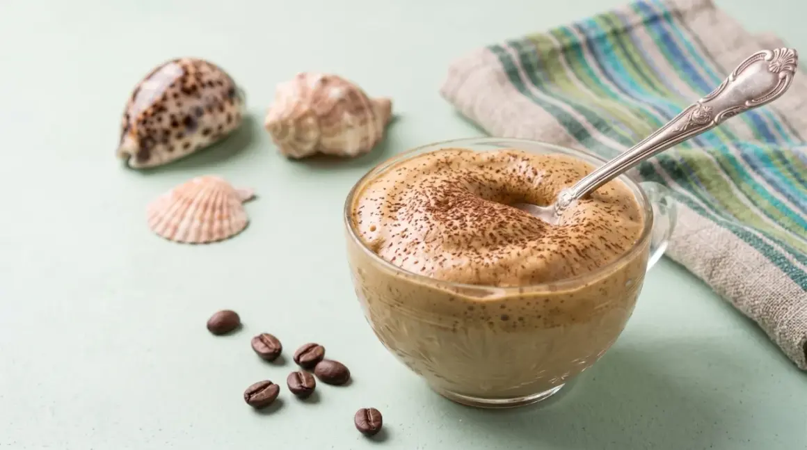 Un cremoso dalgona coffee, spolverato di cacao, accanto a chicchi di caffè e conchiglie.