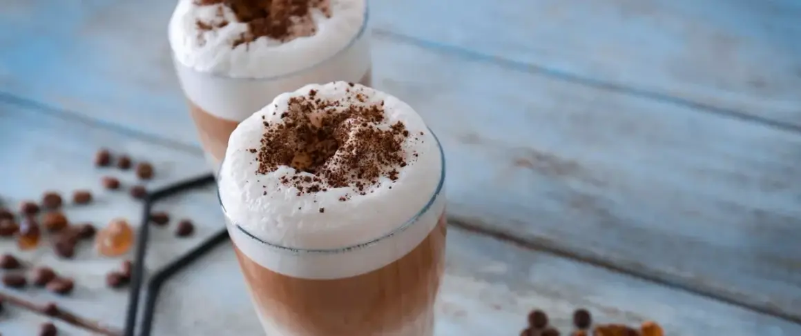 Due bicchieri di latte macchiato freddo con schiuma soffice e cacao in polvere, su un tavolo di legno azzurro.