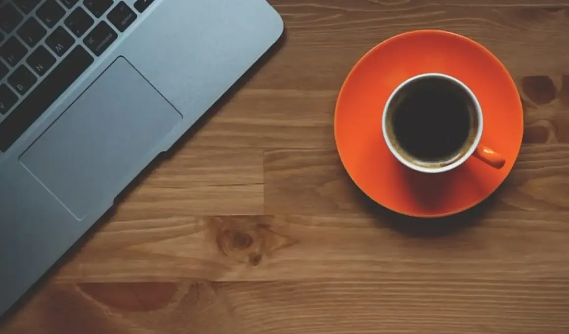 Tazza di caffè nero su piattino arancione accanto a un laptop. Scopri quando bere il caffè per dimagrire.