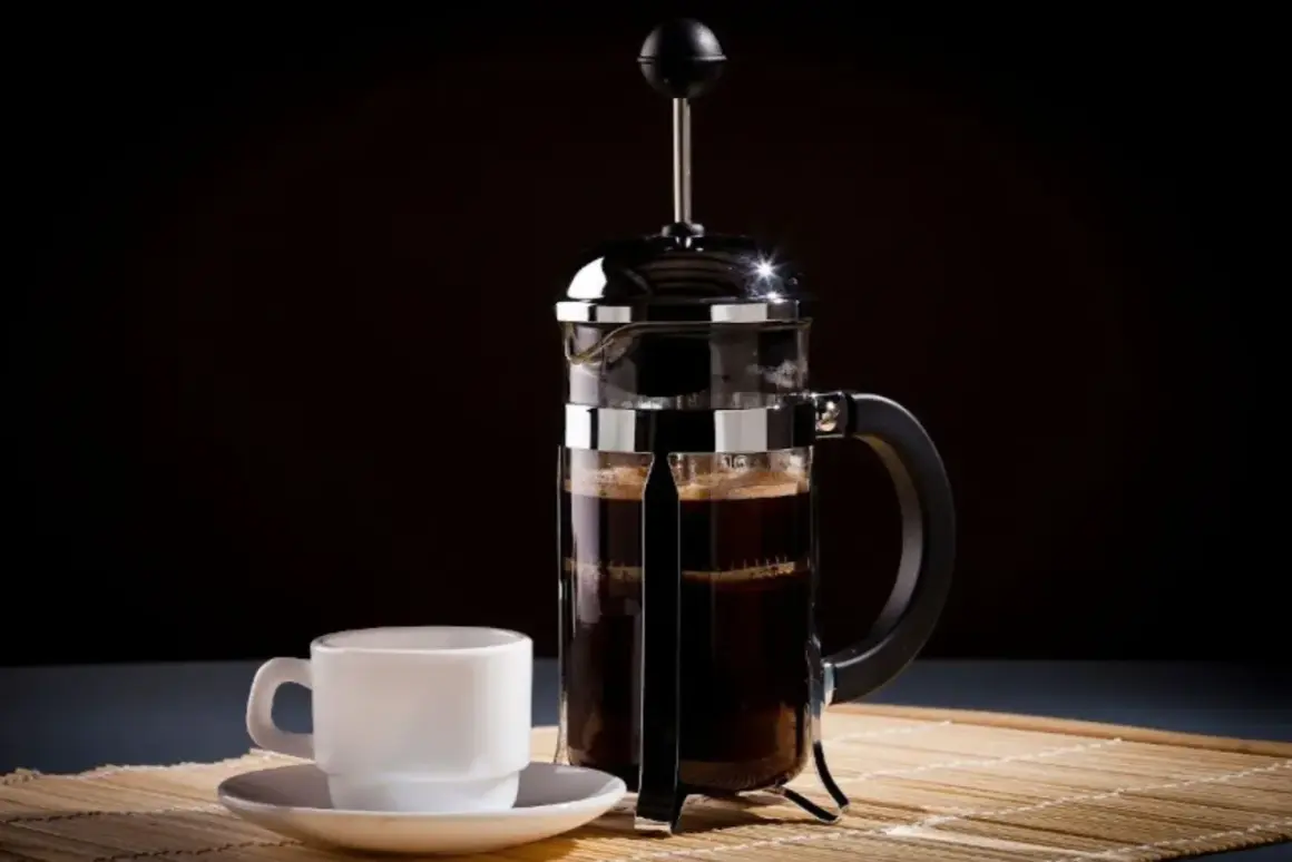 Una tazzina bianca accanto a una caffettiera francese piena di caffè scuro, pronta per essere gustata.