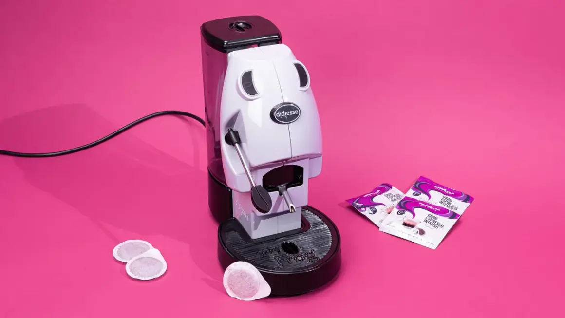Didiesse Frog espresso machine with coffee pods and Lavazza Gran Espresso Intenso packets on a pink background.