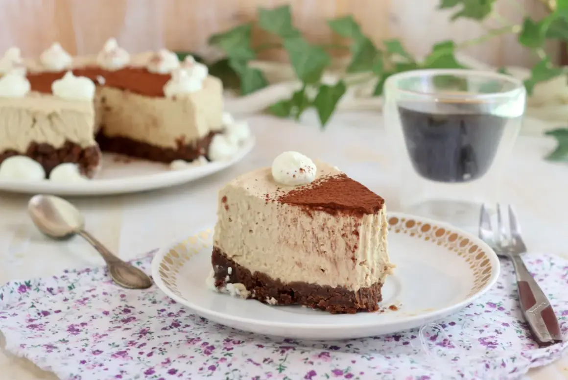 Una fetta di cheesecake al caffè senza gelatina, con base croccante al cacao e decorata con panna e cacao in polvere.