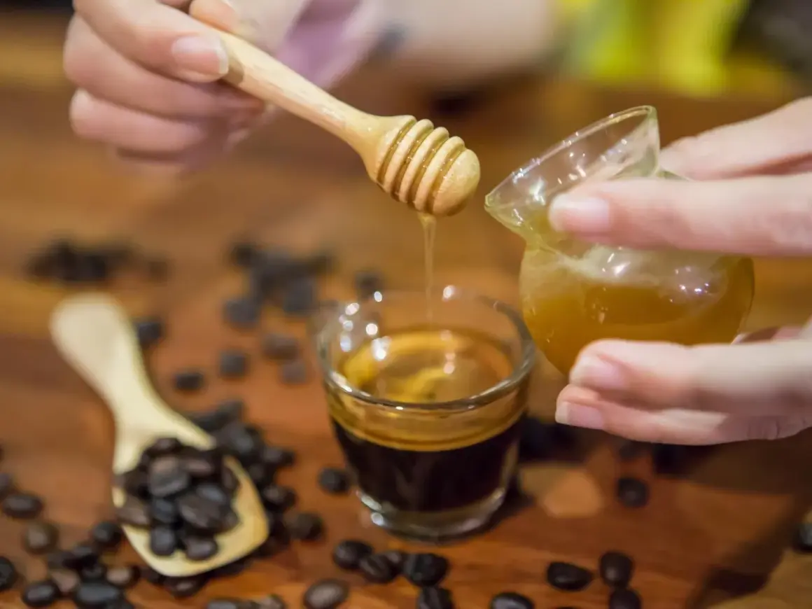 Gocce dorate di miele scendono su un caffè espresso. Chicchi di caffè tostati e un cucchiaino di legno completano la scena.