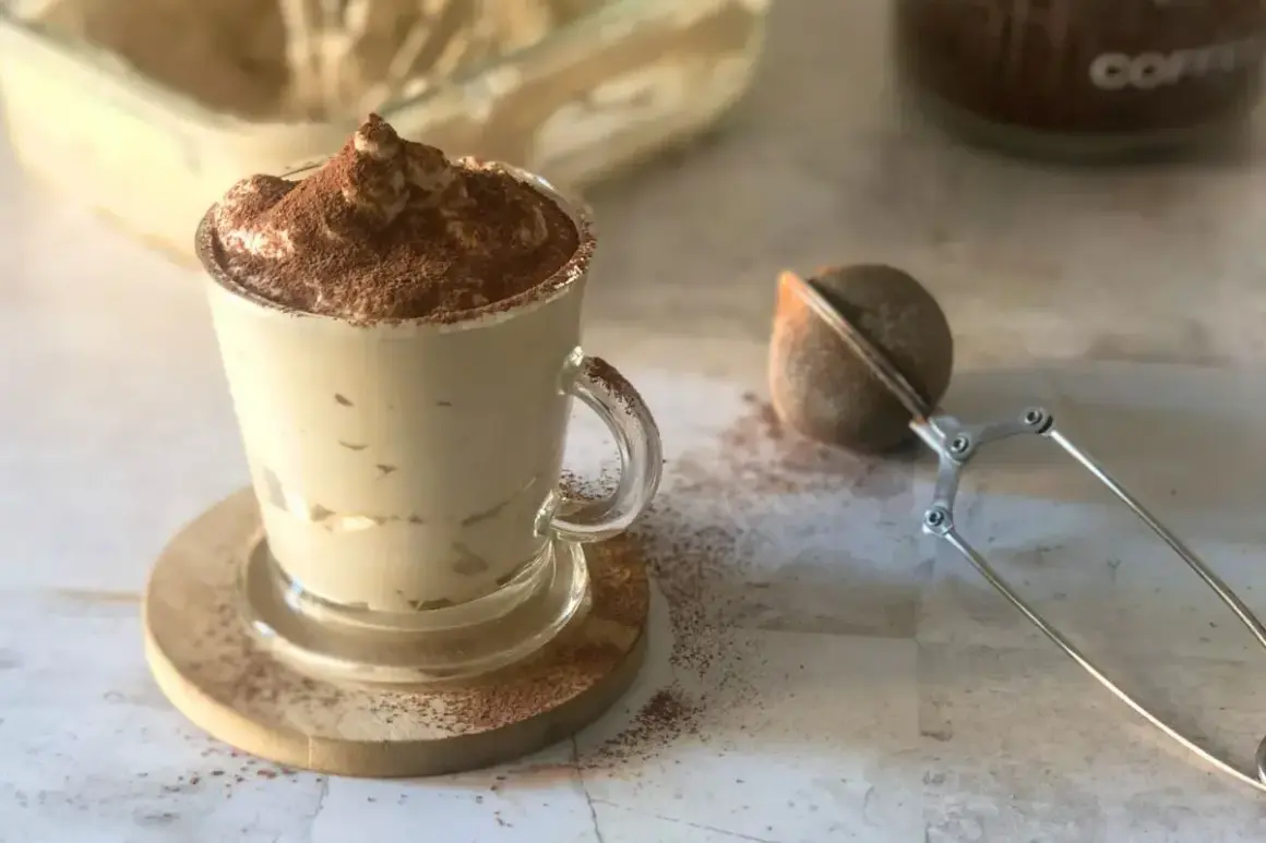 Deliziosa cremina caffè in una tazza di vetro, spolverata di cacao. Accanto, un colino per dolci e un barattolo di caffè.