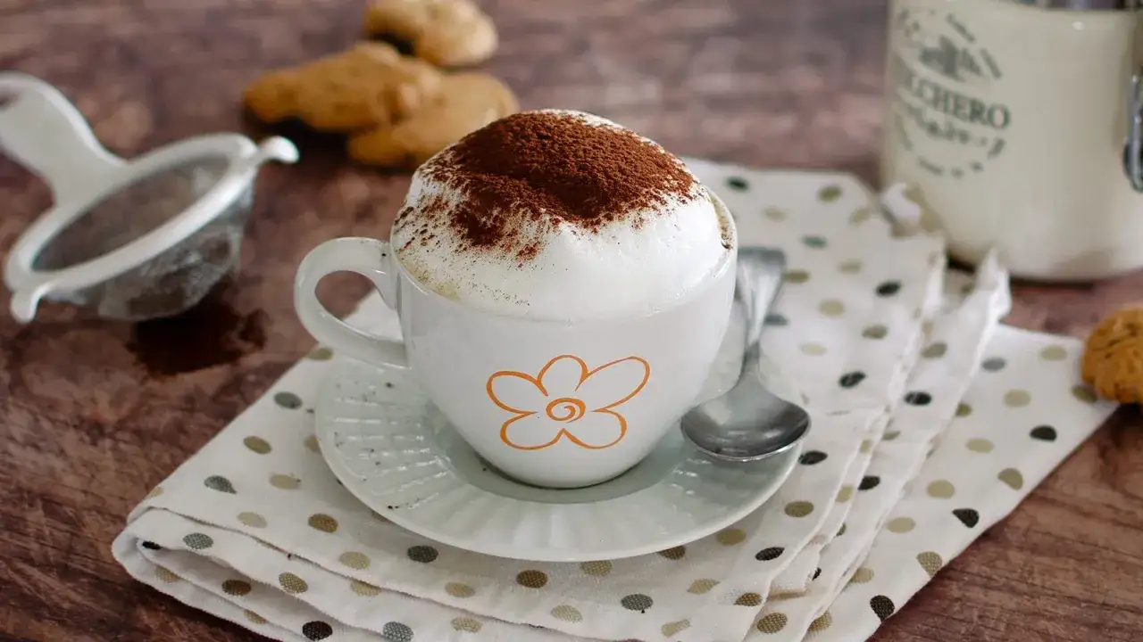 Un invitante cappuccino caldo, spolverato di cacao, pronto per essere gustato con biscotti e un pizzico di zucchero.
