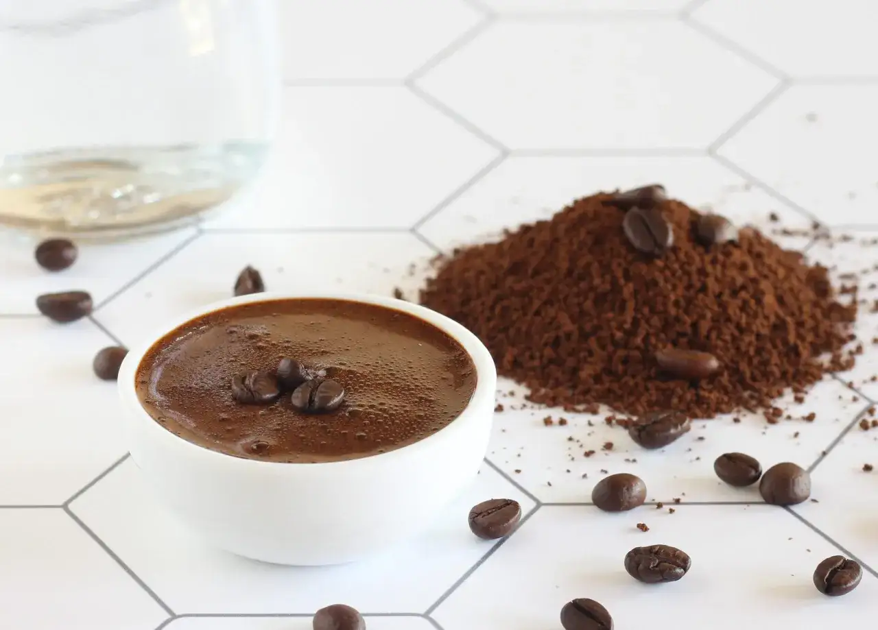 Una tazza di sciroppo al caff&egrave;, chicchi e caff&egrave; macinato sparsi su una superficie bianca con motivo a esagoni.