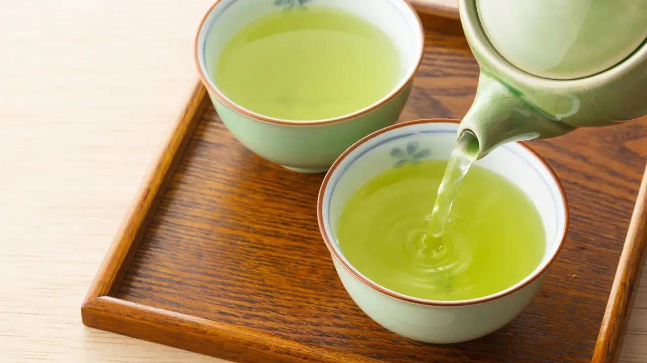 Versando tè verde in una tazza, godendo dei suoi benefici. Due tazze piene di liquido verde brillante su un vassoio di legno.