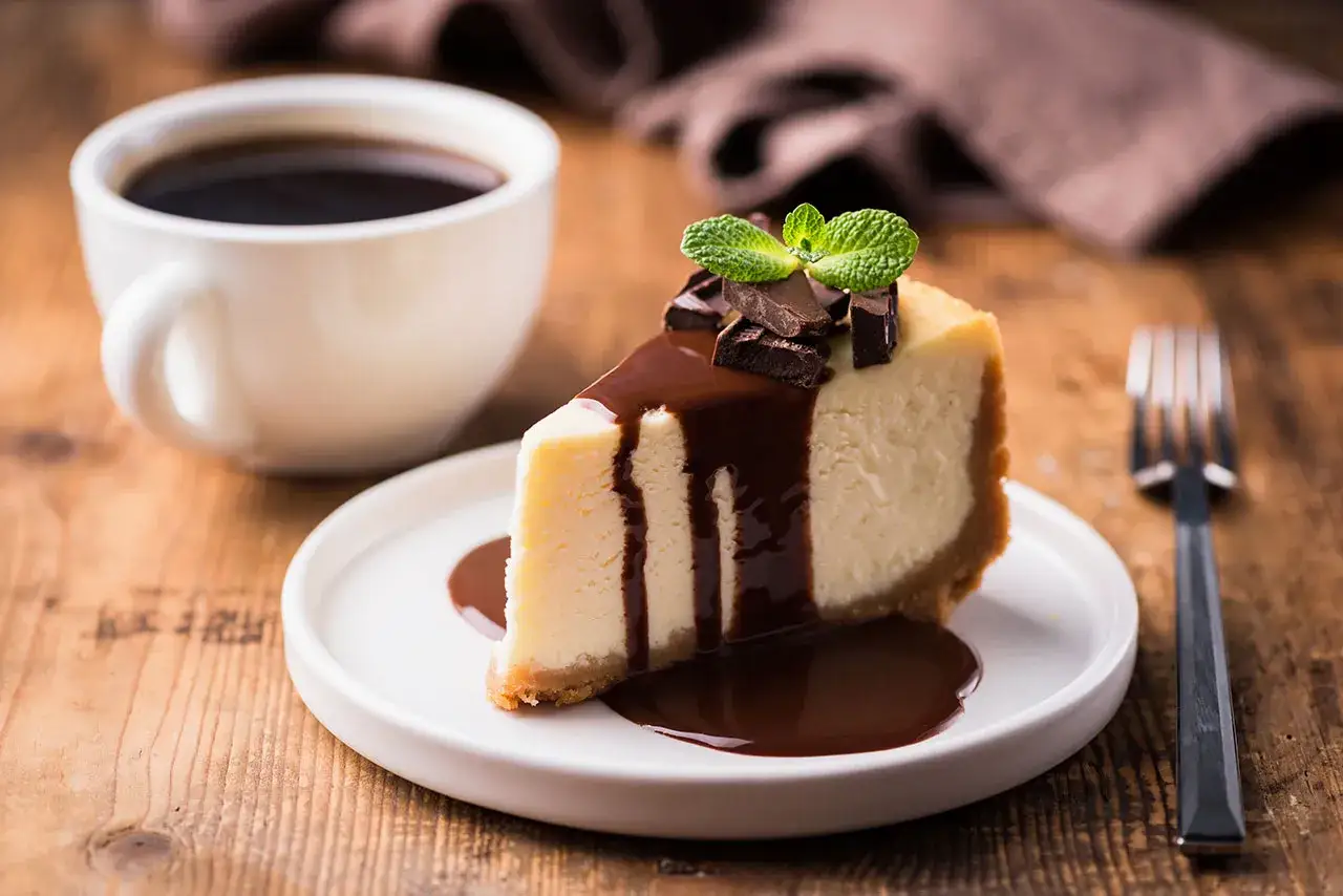 Una fetta di cheesecake al cioccolato e caffè, guarnita con menta e scaglie di cioccolato, su un piatto bianco, accanto a una tazza di caffè fumante.