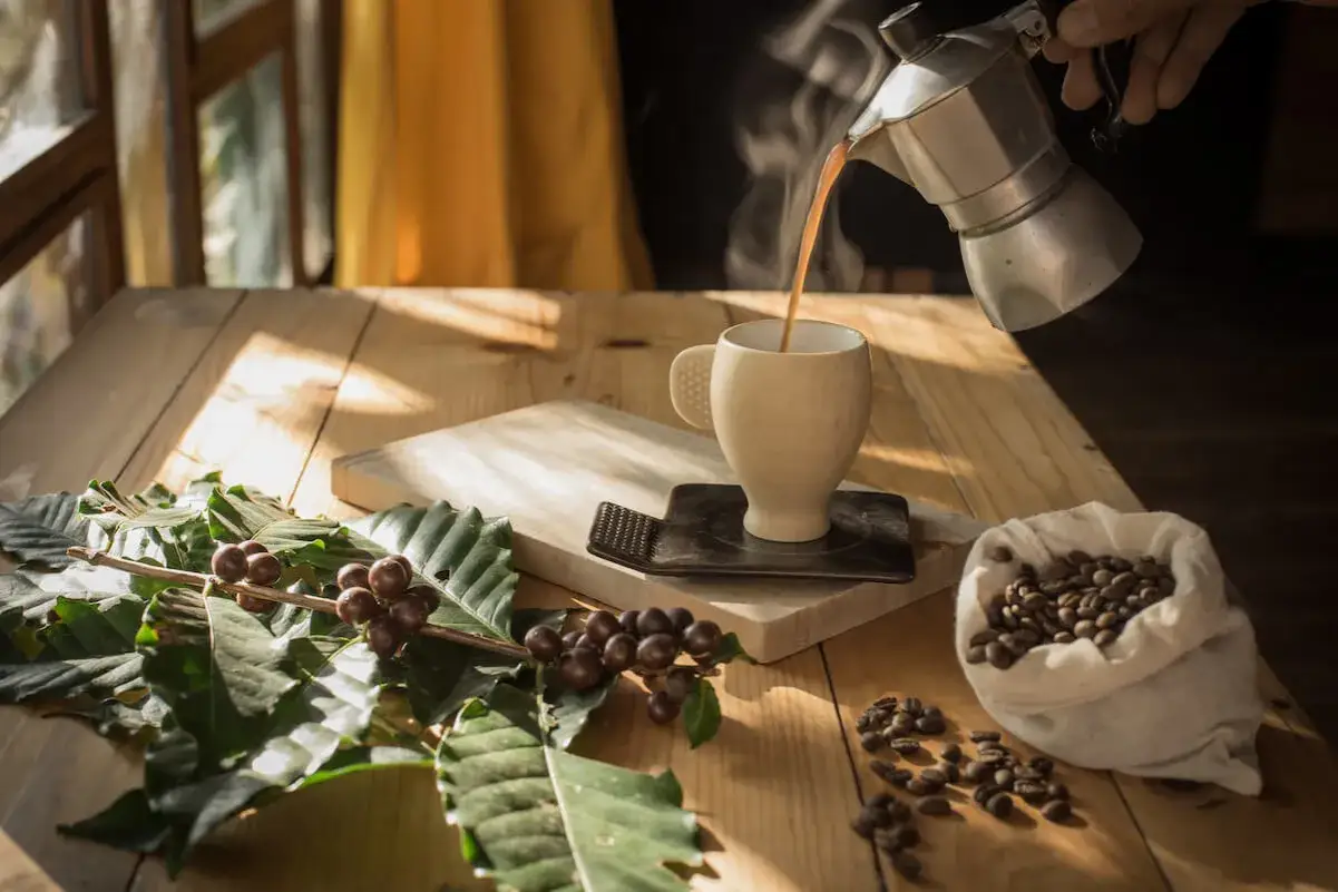 Un filo di caff&egrave; che esce dalla moka riempie una tazza. Sul tavolo, chicchi di caff&egrave; e rami di caff&egrave;.