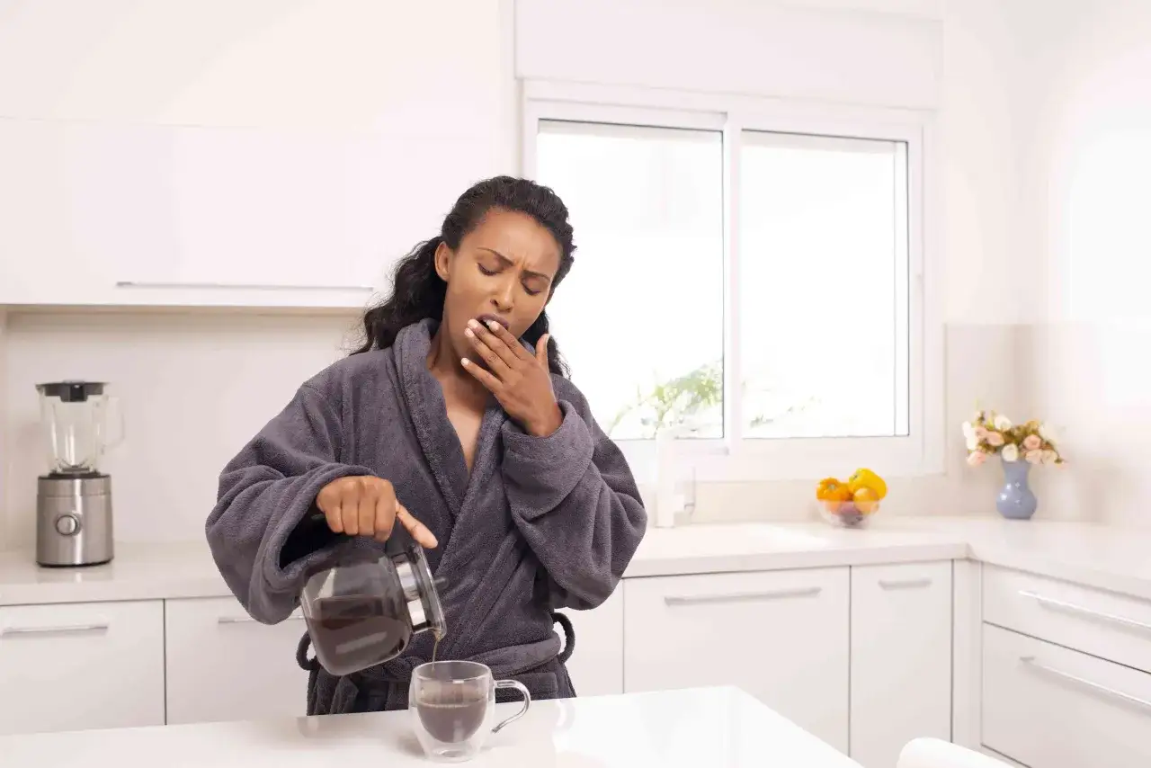 Donna in accappatoio versa caffè, sbadigliando. Potrebbe soffrire di astinenza da caffeina, con sintomi di stanchezza e mal di testa.