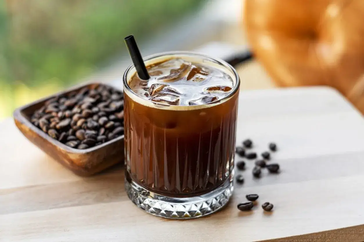 Un bicchiere di cold brew latte rinfrescante con ghiaccio e una cannuccia nera, accanto a chicchi di caff&egrave; tostati in una ciotola di legno.