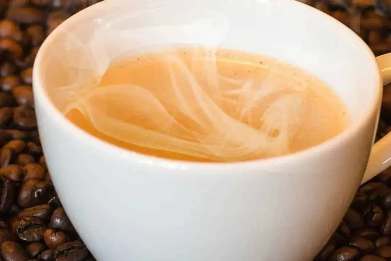 Tazza bianca piena di caff&egrave; fumante, con chicchi di caff&egrave; tostati sullo sfondo. Il vapore crea disegni artistici sulla superficie del caff&egrave; bruciato.