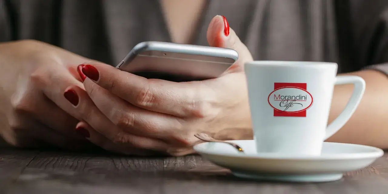 Mani con smalto rosso usano uno smartphone accanto a una tazza di caffè con il logo