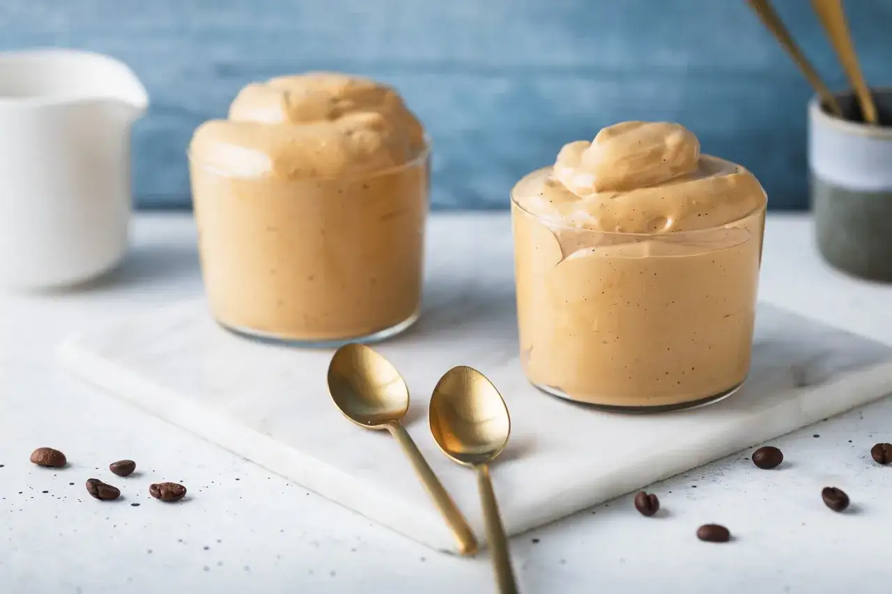 Due bicchieri di cremosa schiuma di caff&egrave;, perfetti per la tua ricetta crema di caff&egrave; senza panna. Due cucchiaini dorati e chicchi di caff&egrave; completano la scena.