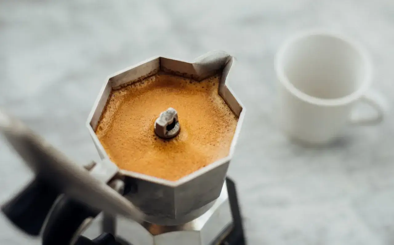 Caffettiera moka piena di caffè fumante, pronta per essere servita in una tazzina bianca. Un invito a bere caffè.